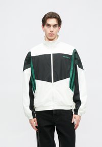 Balts un melns ar rāvējslēdzi aizverama jaka ar zaļiem akcentiem, kurai ir ševronu raksts, elastīgas piedurknes un izšūts logo uz krūšu.