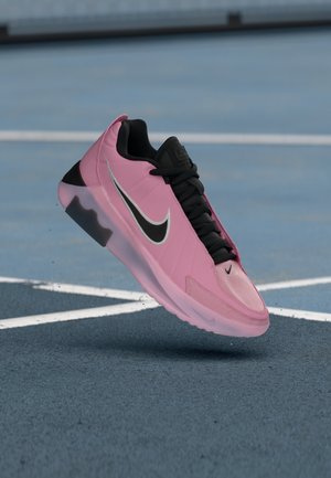 Pink og sort Nike-sneaker med sorte snørebånd, der svæver over en blå sportsbane med hvide linjer.