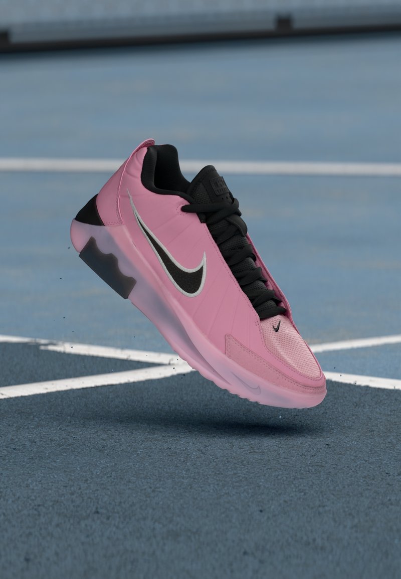 Baskets Nike roses et noires avec des lacets noirs flottant au-dessus d'un terrain de sport bleu avec des lignes blanches.