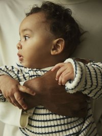 Sweter dzianinowy w paski, kremowy z granatowymi liniami, z prążkowanymi mankietami. Niemowlę trzymane na rękach, z kręconymi włosami i delikatnymi rysami twarzy.