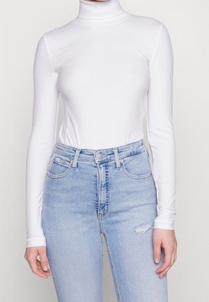 Kobieta ubrana w dopasowany, biały, prążkowany golf z długim rękawem, wpuszczony w jasno-niebieskie, jeansy z wysokim stanem z małym przetarciem na udzie.