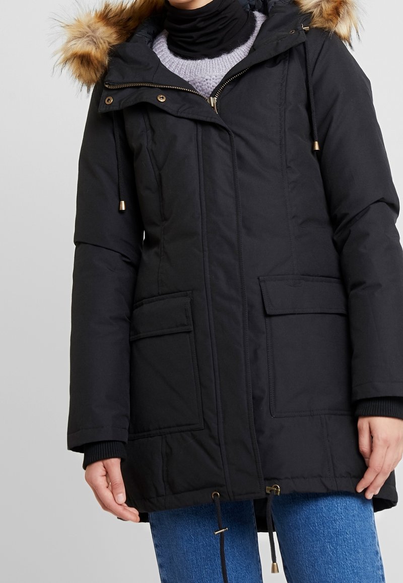 Parka negra, con cremallera en la parte frontal, capucha forrada de piel, dos grandes bolsillos frontales, cordones ajustables en la cintura, tela texturizada.