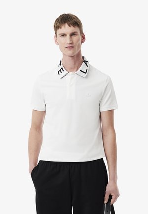 Camisa polo blanca con un cuello de letras negras, mangas cortas y un pequeño logotipo en el pecho. Combinada con pantalones negros y sosteniendo un cinturón.