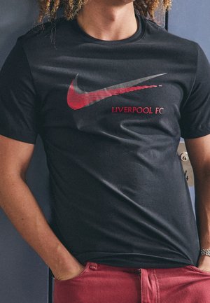 Person trägt ein schwarzes Nike-T-Shirt mit einem roten grafischen Swoosh und dem Schriftzug „Liverpool FC“, kombiniert mit roten Hosen, Hände in den Taschen.