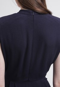 Marinblå klänning med en texturerad tyg, som har en hög halsringning, korta ärmar och en diskret dragkedja i ryggen.