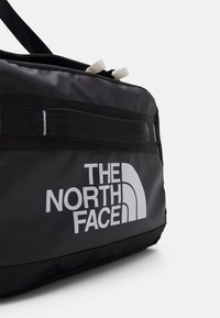 Musta reppulaukku, jossa on The North Face -logo valkoisena, valmistettu kestävästä materiaalista, mustilla nauhoilla ja beigeillä yksityiskohdilla vetoketjun vetimissä.