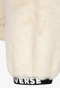 Kremowa kurtka z futra syntetycznego o miękkiej teksturze, z czarnym prążkowanym pasem w talii z białym napisem "UNVERSE". Zaokrąglony, przytulny design.