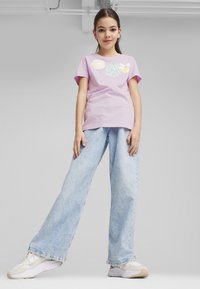 Fioletowy t-shirt z krótkim rękawem z graficznymi detalami, w połączeniu z jasnoniebieskimi spodniami o szerokich nogawkach i białymi trampkami z jasnobeżowymi akcentami.