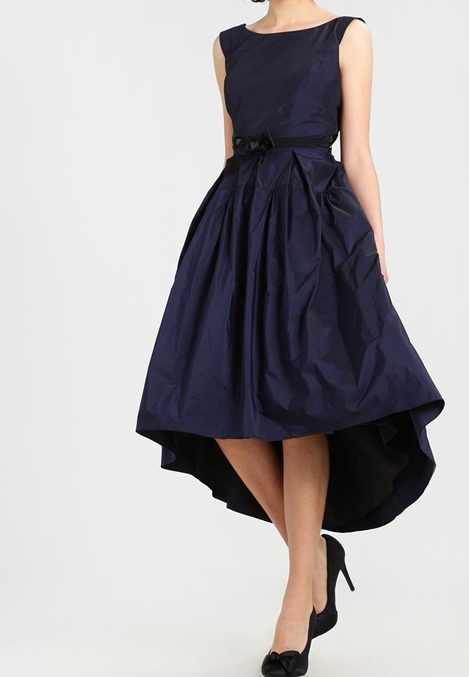 Femme portant une robe sans manches bleu marine à coupe asymétrique avec une petite ceinture nouée et des chaussures à talons noires, debout sur fond blanc.