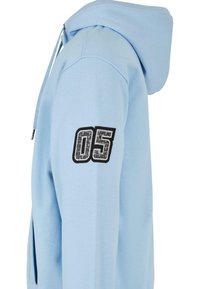 Světlomodrá mikina s kapucí a černou a šedou nášivkou s číslem "05" na horní části rukávu, s jemným vzorovaným designem uvnitř číslic.