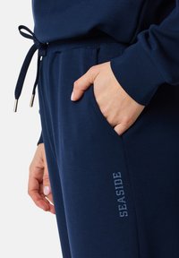 Marine Jogginghosen mit einem Kordelzug-Bund, Seitentasche und dem Aufdruck "SEASIDE" in einem helleren Blau am Oberschenkel. Weicher Stoff.