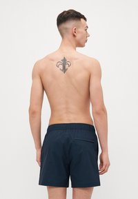 Pantaloni da bagno blu navy con righe verticali, vita elasticizzata, tasca posteriore e una texture liscia. Modello che mostra la vista posteriore.