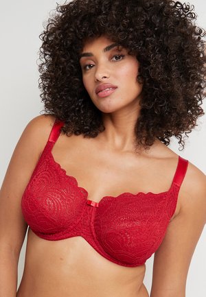 Soutien-gorge en dentelle rouge avec des bords festonnés, doté de bretelles adjustables et d'un petit nœud décoratif au centre. Motif floral texturé sur toute la surface.