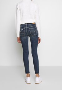 Jeans skinny de color azul oscuro con una cintura de altura media, que presentan un desgaste sutil, costuras intrincadas en los bolsillos y un diseño de pierna ajustada.