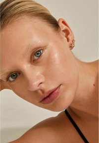 Guld øreringe med flerbladede blomstermotiver prydet med små ædelsten. Glat hudoverflade, neutral baggrund og minimal makeup.