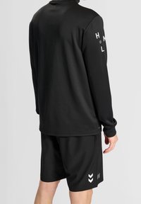 Czarna sportowa koszulka z długim rękawem i okrągłym dekoltem, w zestawie ze spodniami krótki, które mają białe akcenty w kształcie chevron po bokach. Gładka faktura materiału.