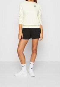Person som bär en krämfärgad Nike-tröja, svarta shorts, vita Nike-strumpor och vita sneakers som står mot en ljus bakgrund.