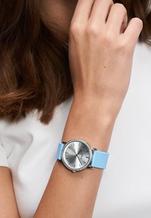 Silberne Armbanduhr mit rundem Zifferblatt, blauem Silikonarmband und Strassakzenten. Das Zifferblatt zeigt ein strukturiertes Muster und römische Ziffern.