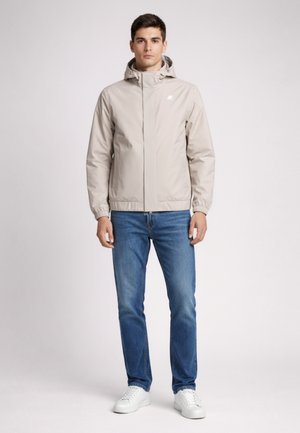 Giovane uomo in piedi, indossa una giacca con cappuccio beige chiaro, jeans blu e sneakers bianche, davanti a uno sfondo neutro.