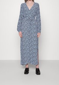 Modrá květinová maxi šaty s dlouhými rukávy, hlubokým výstřihem do V a páskem v pase; mají boční rozparky a hladkou, lehkou tkaninu. V kombinaci s černými dřeváky.