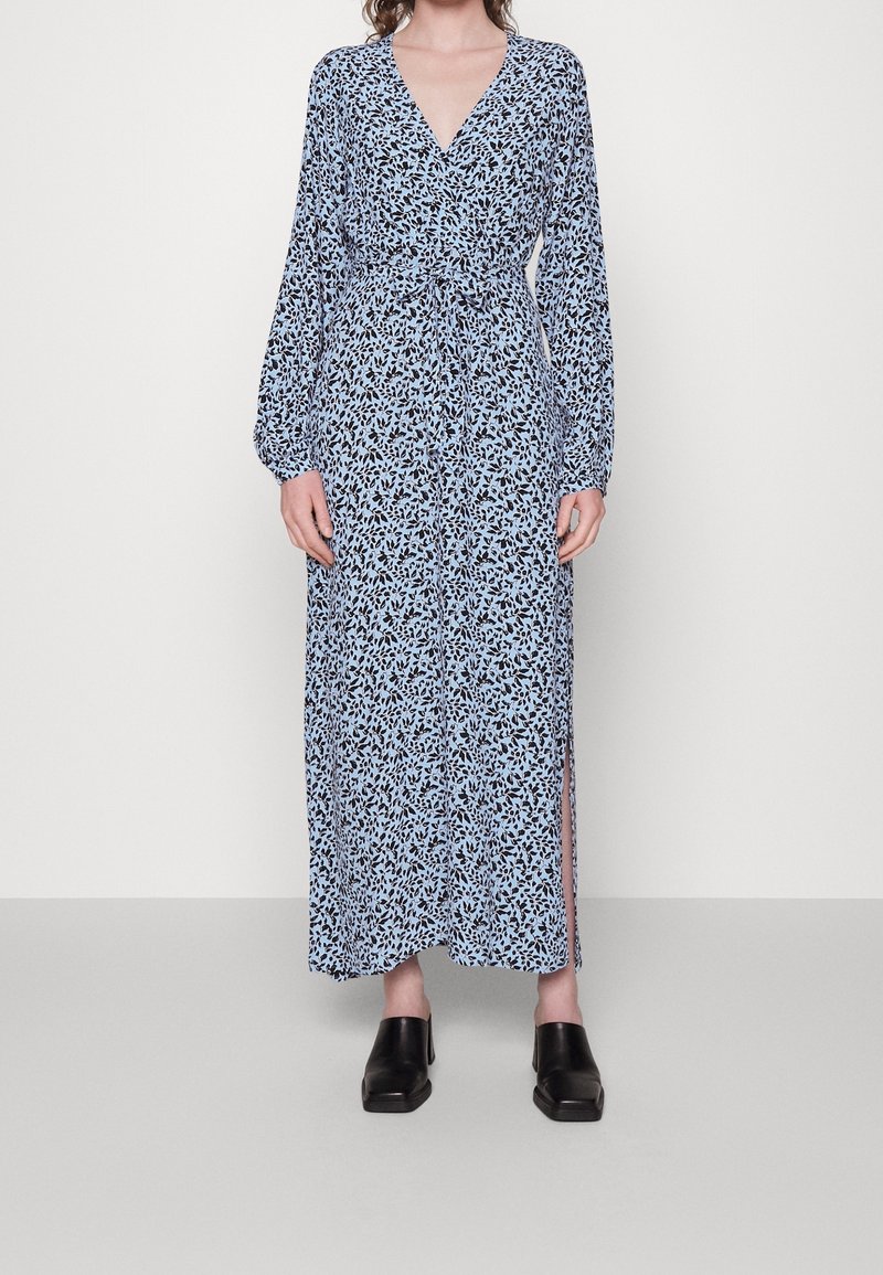 Modrá květinová maxi šaty s dlouhými rukávy, hlubokým výstřihem do V a páskem v pase; mají boční rozparky a hladkou, lehkou tkaninu. V kombinaci s černými dřeváky.