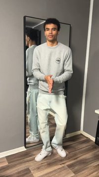 Jeune homme en sweat gris et jean clair debout en train d'applaudir devant un miroir en pied sur un sol en bois à l'intérieur.