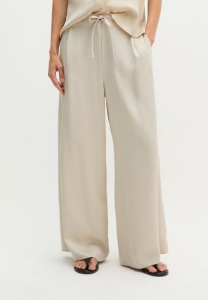 Personne portant un pantalon ample beige à jambes larges avec une taille à cordon et des sandales noires tongs, debout avec une main dans la poche.