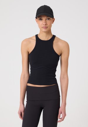 Vrouw draagt een zwarte, mouwloze, nauwsluitende top, legging met hoge taille en een zwarte honkbalpet, staand tegen een lichte achtergrond.