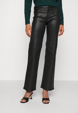 Kobieta w czarnych, skórzanych spodniach z wysokim stanem i prostymi nogawkami, nosząca czarne, otwarte sandały na obcasie oraz ciemnozielony sweter.