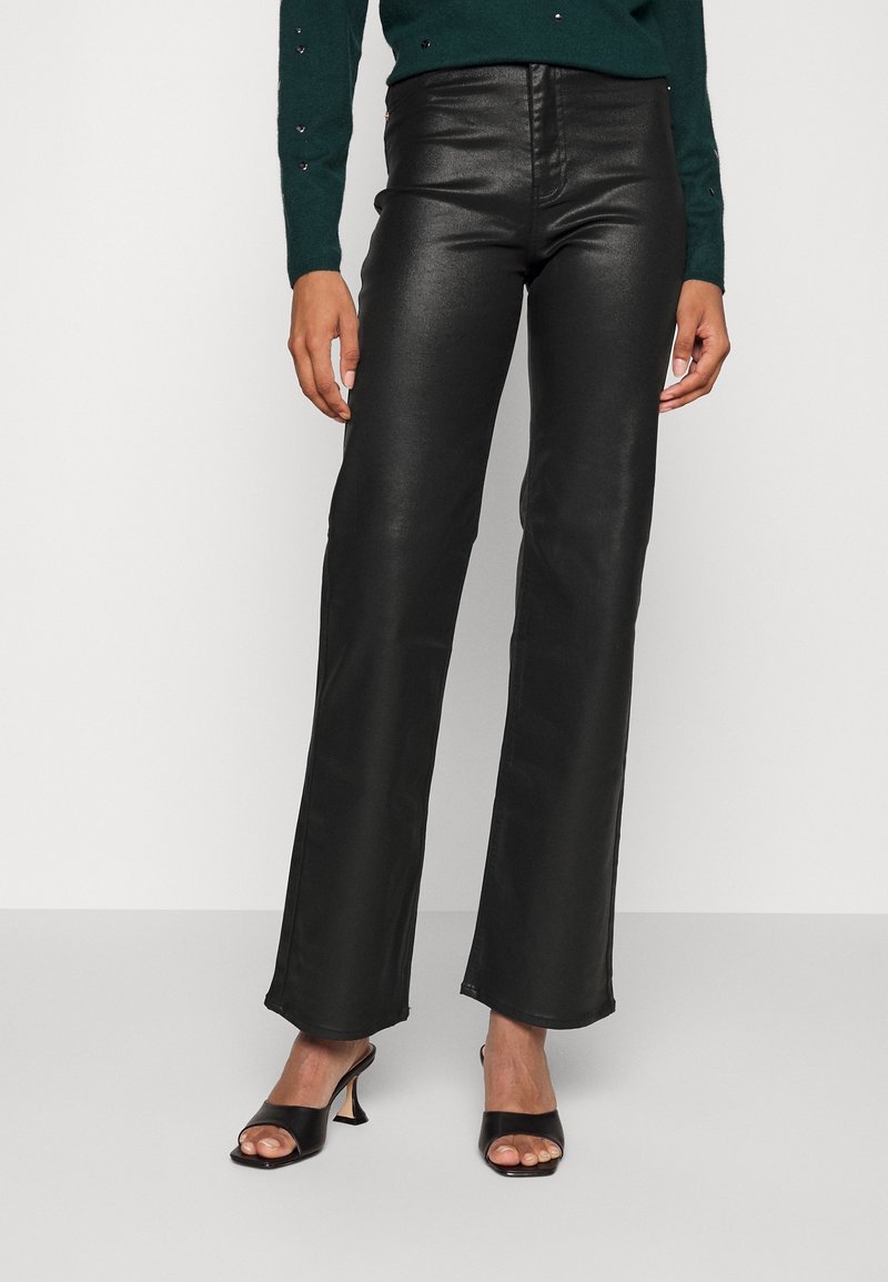 Femme portant un pantalon en cuir noir taille haute à jambe droite, des sandales à talons noires à bouts ouverts et un pull vert foncé.