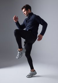 Melnas sporta legginses, tumši zila jaku ar rāvējslēdzēju un sudraba skriešanas apavi ar tīkla augšdaļu. Modelis kustībā, ceļot vienu ceļgalu.
