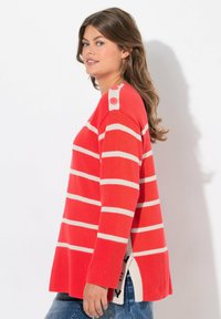 Czerwony sweter w paski z kremowymi poziomymi paskami, żebrowana tekstura, rozcięcia po bokach oraz akcenty z guzikami na ramieniu. Luźny fason i design.