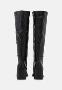 Bottes noires montantes au-dessus du genou en cuir lisse avec un talon épais et une fermeture éclair à l'arrière. Présentent une silhouette ajustée et un design minimaliste.