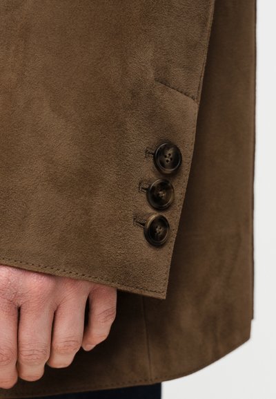 Gros plan sur la manche d'une veste en daim marron avec trois boutons foncés et une main partiellement visible sous le poignet.