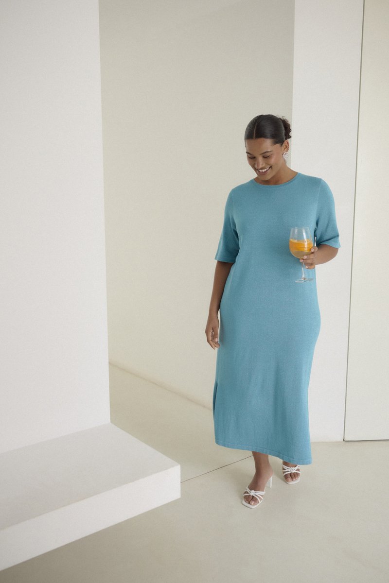 Turkis, knælang kjole med korte ærmer og sideslids, lavet af blødt materiale. Person holder et glas med en orange drik og har sandaler på.