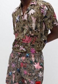Mann, der ein passendes braunes, floral bedrucktes Kurzarm-Hemd mit Knopfleiste und Shorts mit Kordelzug in Rosa-, Lila- und Grüntönen trägt.