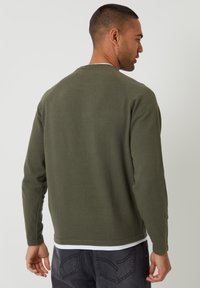 Olijfgroene gebreide trui met lange mouwen, ronde hals en geribbelde manchetten, gecombineerd met grijze spijkerbroek. Achteraanzicht toont een gladde textuur.