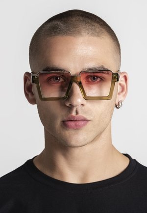 Ung mand med overskåret kort hår iført store firkantede solbriller med lyserøde linser og en sort skjorte, vendt fremad mod en ensfarvet baggrund.