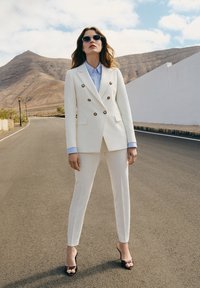 Biała marynarka dwurzędowa z złotymi guzikami, zestawiona z dopasowanymi, wąskimi spodniami i jasnoniebieską koszulą z kołnierzykiem. Czarne okulary przeciwsłoneczne i szpilki.