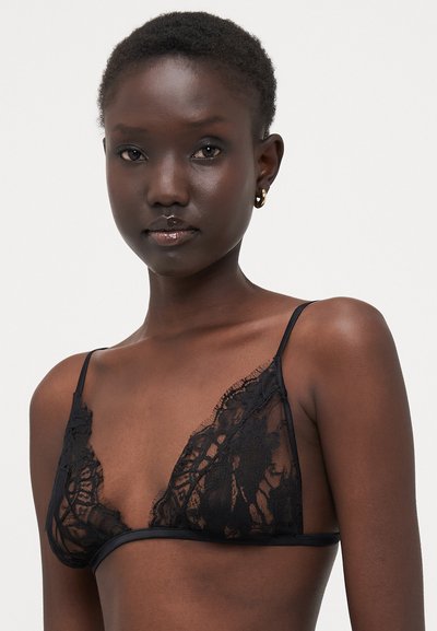 Bralette de encaje negro que presenta una forma triangular con tejido transparente, tirantes delgados ajustables y patrones florales en las copas.
