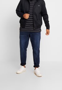 Man med svart jacka, mörkgrå och svart randig skjorta, uppvikt mörkblå jeans och vita sneakers som står på vit golv.