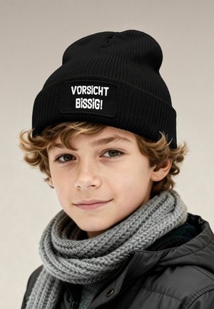 Black knit beanie with a white rectangular patch reading "VORSICHT BISSIG!" Grey knitted scarf and black jacket with a smooth texture.
