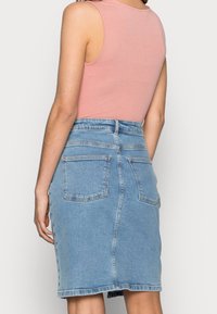 Donna che indossa un top senza maniche rosa infilato in una gonna di jeans azzurro chiaro a vita alta con tasche posteriori, vista da dietro.