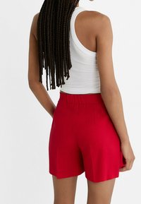 Shorts en lin rouges avec une taille élastique, coupe ajustée et poches, visibles de dos avec un débardeur blanc et des cheveux tressés.
