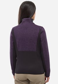 Femme vue de dos portant une veste violette et noire avec un col montant et un pantalon beige sur fond blanc.