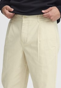 Pantaloni plisati bej dintr-un material ușor. Au închidere cu nasture și buzunare laterale. Textură netedă și croială dreaptă a picioarelor.