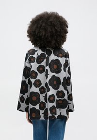 Person with curly hair wearing a gray jacket featuring large black and brown floral patterns, paired with blue jeans, seen from behind.