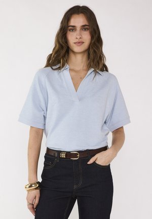 Vrouw met golvend bruin haar draagt een lichtblauw, kort mouwtjes, overhemd met kraag dat in donkere jeans met een bruine riem is gestopt, goudkleurige armbanden en ringen.