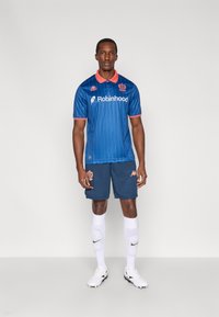 Mėlyna futbolo marškinėliai su vertikaliomis juostelėmis, oranžine apykakle ir logotipu. Derinami su tamsiomis šortais ir baltais kojinėmis. Juodi futbolo bateliai.