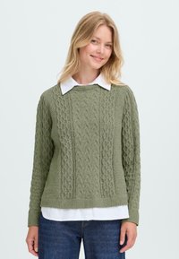 Groene gebreide trui met een gestructureerd patroon, ronde halslijn en geribbelde manchet, gedragen over een witte overhemd met kraag.